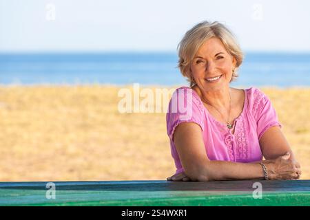 Porträt einer attraktiven, eleganten Seniorin, die draußen sitzt, mit Strand und Meer hinter ihr. Stockfoto