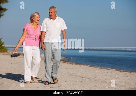 Ein glücklicher, lächelnder älterer Mann und eine Frau, das alte pensionierte Paar, das am Strand spaziert, lacht und Hände hält Stockfoto