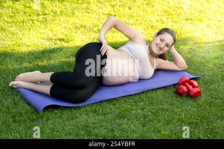 Glückliche lächelnde schwangere Frau, die auf einer Fitnessmatte im Park trainiert Stockfoto