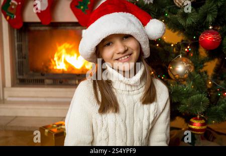 Porträt eines 10-jährigen Mädchens mit Weihnachtsmann-Hut, der neben dem Kamin und dem geschmückten Weihnachtsbaum sitzt Stockfoto