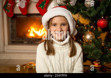 10-jähriges Mädchen mit Weihnachtsmann-Hut, das neben dem Kamin sitzt und den Weihnachtsbaum dekoriert hat Stockfoto