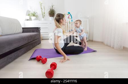 Schöne lächelnde Mutter mit einem Kleinkind, der Yoga auf einer Fitnessmatte im Wohnzimmer praktiziert Stockfoto