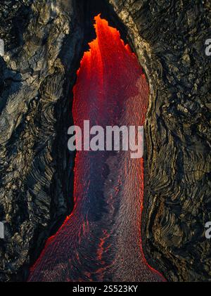 Aus der Vogelperspektive pulsierende geschmolzene Lava, die durch eine vulkanische Spalte fließt, im Kontrast zu dunklem vulkanischem Gestein in einer dramatischen Landschaft. Stockfoto