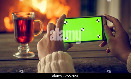Getöntes Nahaufnahme Foto von weiblichen Händen, die auf dem Smartphone Fotos von Tee und brennendem Kamin in der Nacht machen. Leerer grüner Bildschirm zum Einfügen eines eigenen Bildschirms Stockfoto