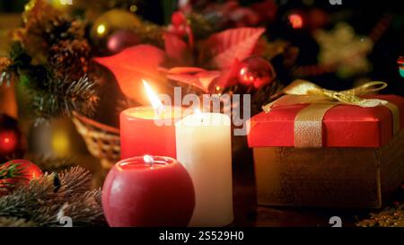 Nahaufnahme von drei brennenden Kerzen und goldener Geschenkbox mit Schleife. Perfekte Kulisse für Winterferien und Feiern. Nahaufnahme von Stockfoto