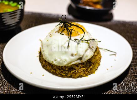 koreanische Küche: Kimchi Bokkeum bap (gebratener Reis mit Kimchi, Rindfleisch und Spiegelei) auf weißem Teller auf braunem Lederbrett im lokalen Restaurant Stockfoto