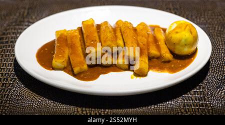 Die koreanische Küche - Tteok bokki Vorspeise (Reis Sticks mit gekochtem Ei in pikanter Sauce) auf die weiße Platte auf Braun leathed Board in einem lokalen Restaurant Stockfoto