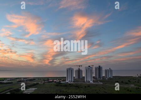Faszinierende Farbtöne erfüllen den Abendhimmel, während Kraniche über modernen Gebäuden in der Nähe der ruhigen Küste aufragen. Stockfoto