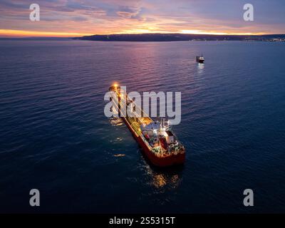 Aus der Vogelperspektive eines hell beleuchteten Tankschiffes auf dem Meer während der Dämmerung, umgeben von offenem Wasser und einem leuchtenden Horizont Stockfoto