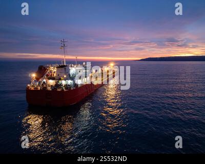Aus der Vogelperspektive eines hell beleuchteten Tankschiffes auf dem Meer während der Dämmerung, umgeben von offenem Wasser und einem leuchtenden Horizont Stockfoto