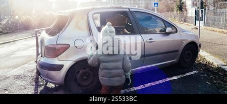 Ein Kind in einem warmen Mantel und Hut, das in der Nähe eines beschädigten und verlassenen silbernen Autos steht, in einem urbanen Außenbereich mit Sonnenlicht in Frankreich Stockfoto