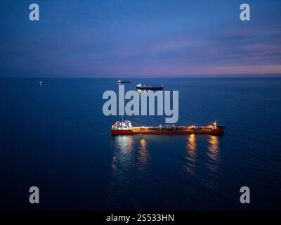 Aus der Vogelperspektive eines hell beleuchteten Tankschiffes auf dem Meer während der Dämmerung, umgeben von offenem Wasser und einem leuchtenden Horizont Stockfoto