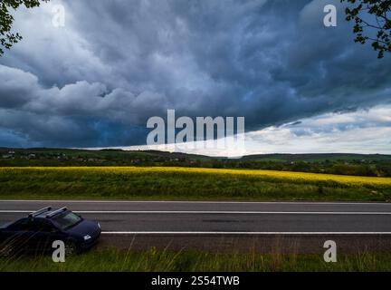 Frühlingsgelb blühende Rapsfelder, regionale Straße, dramatisch bewölktes Gewitter, regnerischer Himmel und ländliche Hügel. Natürliches saisonales Klima, Stockfoto