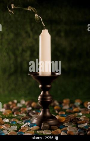 Einzelne weiße Kerze mit Rauchspur auf einem schwarzen Metallkerzenhalter, umgeben von bunten Felsen und Kieselsteinen Stockfoto
