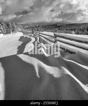 Graustufen. Malerische schwingende Schatten auf Schnee vom Holzzaun. Alpines Bergdorf am Rande, verschneite Wege, Tannenwald. Hochauflösendes Bild Stockfoto