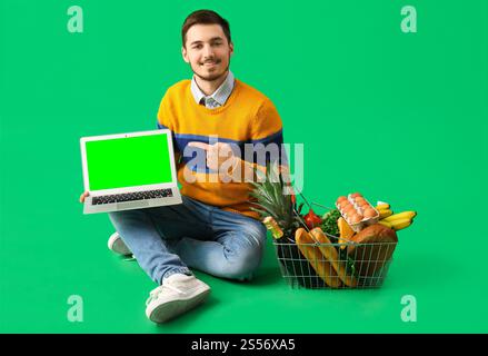 Junger Mann mit Warenkorb, der auf Laptop zeigt, mit leerem Bildschirm auf grünem Hintergrund. Modell für Design Stockfoto