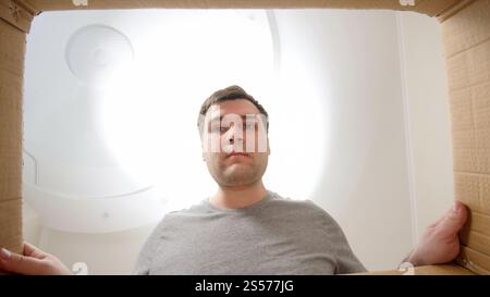 Porträt eines jungen Mannes, der nach dem Umziehen in einen Karton mit Päckchen oder Dingen schaut. Porträt eines jungen Mannes, der in Pappkarton mit Päckchen schaut Stockfoto