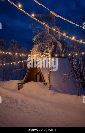 Schneebedeckte Kabine mit A-Rahmen, beleuchtet durch Lichterketten in einem Winterwunderland, schafft eine warme und einladende Atmosphäre unter einem dunkelblauen Nachthimmel Stockfoto