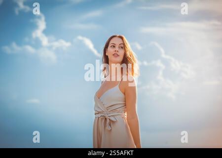Junge Brünette, die steht, trägt ein cremeweißes Kleid mit leicht bewölktem Himmel Stockfoto