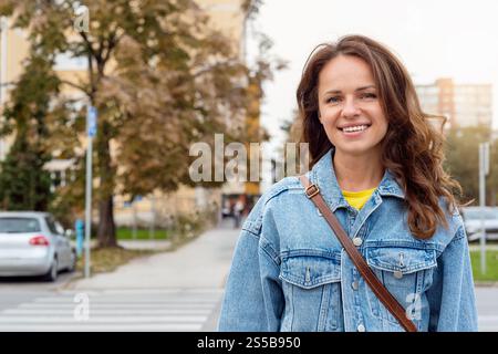 Porträt einer jungen brünetten Frau in Jeansjacke, die auf der Straße steht und in die Kamera blickt und lächelt. Stockfoto