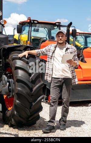Kaukasischer Erwachsener mit digitalem Tablet in der Hand steht neben einem Bau- oder landwirtschaftlichen Traktor. Stockfoto