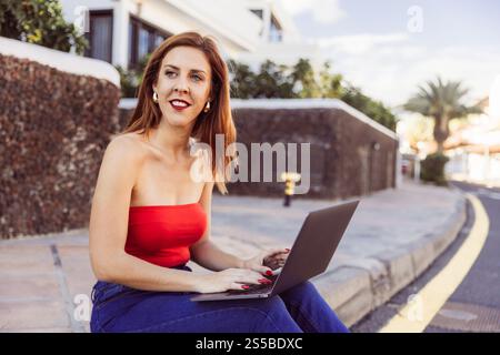 Eine Frau in einem roten Oberteil sitzt auf einem Bürgersteig mit einem Laptop vor sich. Sie konzentriert sich auf ihre Arbeit, möglicherweise Tippen oder Surfen im Internet. Konzept o Stockfoto
