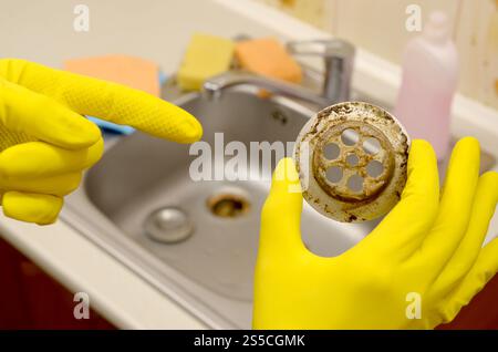 Reiniger in Gummihandschuhen zeigt Abfall im Steckdosenschutz eines Küchenwaschbecken. Verschmutzter Ablassfilter aus Stahl. Abfallfalle Gitter der Entwässerungslöcher. Stockfoto
