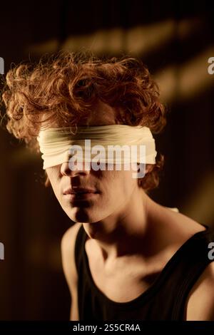 Vertikales Hochwinkelporträt eines lockigen rothaarigen Mannes mit weißem Verband an Augen, der vor der Kamera sitzt Stockfoto