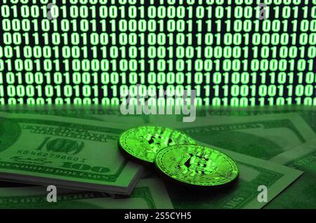 Zwei Bitcoins liegen auf einem Stapel von Dollarscheinen auf dem Hintergrund eines Monitors, der einen Binärcode aus hellgrünen Nullen und eine Einheit auf einem schwarzen zeigt Stockfoto