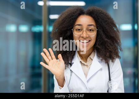Online-Videoberatung, junge, erfolgreiche Ärztin schaut in die Kamera und winkt mit der Hand, kommuniziert aus der Ferne mit Patienten des Kunden. Webcam-Ansicht, Frau im weißen Mantel, mit Stethoskop. Stockfoto