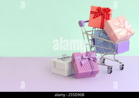 Kleine Geschenkboxen in verschiedenen Farben mit Bändern liegen im Warenkorb auf einem violetten und blauen pastellfarbenen Hintergrund. Kleine Geschenkboxen in verschiedenen Farben Stockfoto