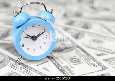 Zeitmanagement-Konzept. Blauer Wecker liegt auf einer großen Menge von hundert Dollar Scheinen. Zeit als eine der wichtigsten Ressourcen im Unternehmen. Zeit als ein Stockfoto