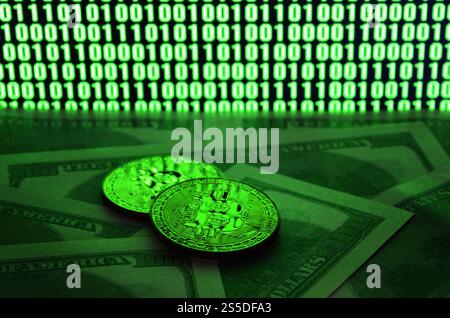 Zwei Bitcoins liegen auf einem Stapel von Dollarscheinen auf dem Hintergrund eines Monitors, der einen Binärcode aus hellgrünen Nullen und eine Einheit auf einem schwarzen zeigt Stockfoto