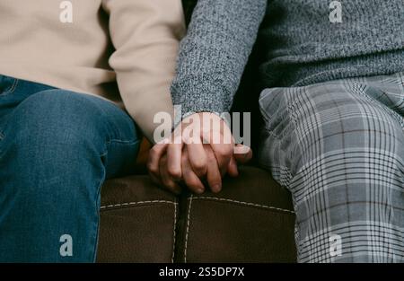 Nahaufnahme von zwei Männern, die legere Kleidung tragen, Hände halten, mit gefalteten Händen auf einem braunen Sofa sitzen Stockfoto