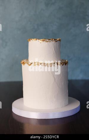 Weiße zweistufige Hochzeitstorte, die mit Blattgold verziert ist, steht auf einem weißen Ständer auf einem dunkelbraunen Tisch vor blauem Hintergrund Stockfoto