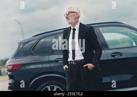 Selbstbewusster Geschäftsmann, der in der Nähe des Autos auf dem Parkplatz in der Stadt stand, Hände in den Taschen, trug einen formellen Anzug Stockfoto