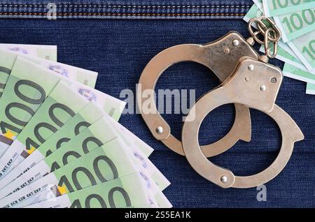 Der Fan von vielen Euro-Scheinen und Polizeihandschellen ist auf einer dunklen Jeansfläche. Hintergrundbild Stockfoto