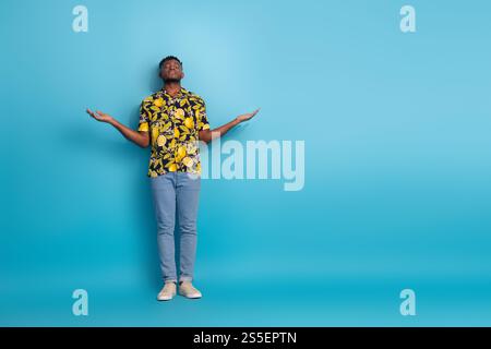 Fröhlicher junger Mann in stilvollem Hemd trifft eine fröhliche Pose vor blauem Hintergrund und verkörpert Sommer-Vibes und Glück Stockfoto