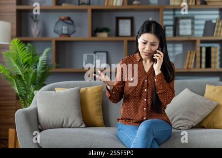 Besorgte asiatische Frau, die ihr Smartphone benutzt, auf dem Sofa sitzt und wichtige Themen diskutiert. Zeigt Emotionen, Kommunikation und Technologie in einem modernen Wohnzimmer. Stockfoto