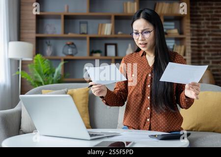 Asiatische Frau, die zu Hause Papiere analysiert, Brille trägt, Laptop benutzt. Konzentration auf wichtige Details, Multitasking im Wohnzimmer. Konzept von Konzentration, Home Office, Papierkram. Stockfoto