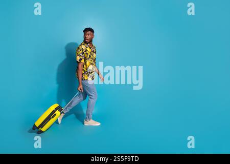 Junger Mann in stilvollem Hemd mit Print und Koffer auf leuchtendem blauem Hintergrund bereit für Reise-Abenteuer Stockfoto