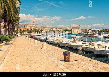 Sonniger Tag an einem mediterranen Yachthafen mit verankerten Booten und einer von Bäumen gesäumten Promenade, die zu historischen Gebäuden in Split, Kroatien, führt Stockfoto