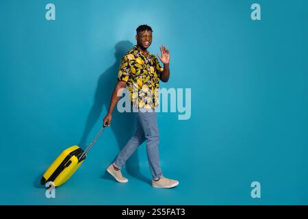 Glücklicher junger Mann, der mit gelbem Koffer in stilvollem Hemd winkt und auf blauem Hintergrund steht Stockfoto
