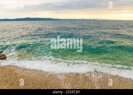 Die Wellen brechen sanft an einem Kiesstrand an der Adria mit einer ruhigen Meereslandschaft und bewölktem Himmel in der Abenddämmerung, Podgora, Kroatien Stockfoto