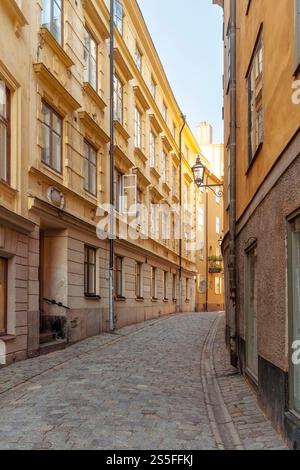 Enge Kopfsteinpflasterstraße, die sich bei Sonnenuntergang zwischen traditionellen alten europäischen Gebäuden schlängelt, mit warmem Licht, das von den Fassaden in der Altstadt von Gamla Stan in Stockholm, Schweden, reflektiert wird Stockfoto