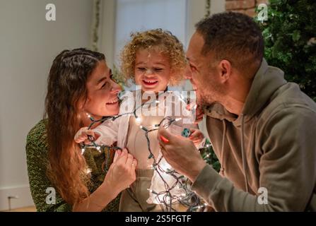 Eine Familie genießt während der Weihnachtszeit schöne gemeinsame Zeit mit einem Kleinkind, das in leuchtende Weihnachtslichter gehüllt ist und zwischen seinen fröhlichen Eltern in Ohio, USA lächelt Stockfoto
