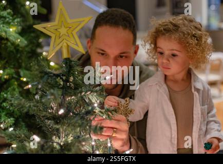 Vater und Tochter des Kleinkindes schmücken einen Weihnachtsbaum mit Lichtern und einem Sternenhimmel zu Hause in Ohio, USA Stockfoto