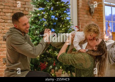 Die Familie genießt einen verspielten Moment in der Nähe eines Weihnachtsbaums, während eine Mutter ein Kind küsst, das von einem lächelnden Vater gehalten wird, Ohio, USA Stockfoto