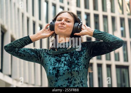Lächelnde Frau in einem blau gemusterten Top genießt Musik auf großen schwarzen Kopfhörern draußen mit Gebäuden im Hintergrund, Berlin, Deutschland Stockfoto