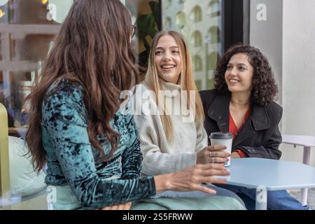 Drei Frauen unterhalten sich an einem Café-Tisch, eine hält eine Kaffeetasse und lächelt hell, Berlin Stockfoto